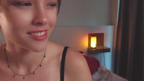 Media: Video of a smiling woman with light skin, wearing a black strapless top, necklace, and earrings. Background features a lit lamp and dark curtains in a dimly lit room.