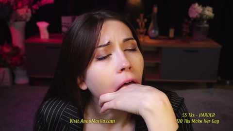 Media: Video of a young woman with long dark hair, wearing a black pinstriped shirt, lying on a carpeted floor, hand near her mouth, eyes closed, in a dimly lit room.