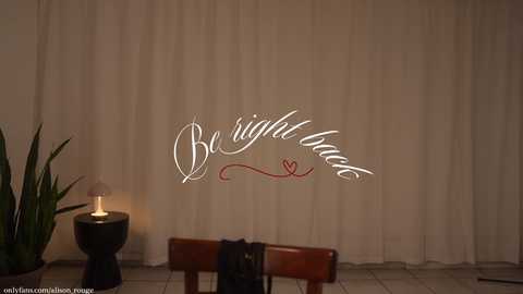 Media: Video of a serene indoor scene with a beige curtain backdrop. Central text reads \"Be right back\" in elegant script, accompanied by a heart symbol. A lit candle on a black stand and a wooden bench are visible in the foreground.