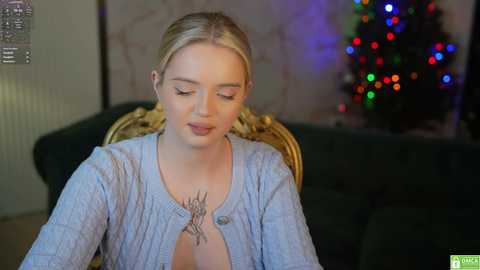 Media: Video of a fair-skinned, blonde woman with closed eyes, wearing a light blue cardigan, seated in a dark green chair. She has a tattoo on her chest. A festive Christmas tree with colorful lights is in the background.