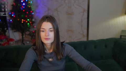 Media: Video of a young woman with straight brown hair, fair skin, and a slender build, wearing a gray long-sleeve top, seated on a green velvet couch in a cozy living room with a lit Christmas tree and festive lights in the background.