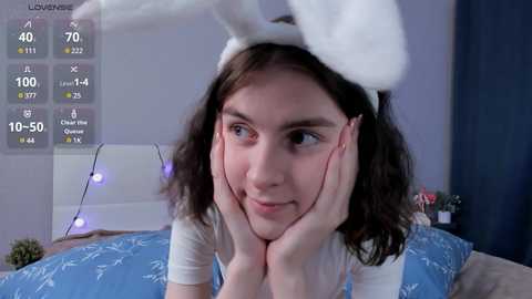 Media: Video of a young woman with fair skin, wearing a white bunny costume with fluffy ears, and lying on a bed with blue sheets. She has a surprised expression and rests her chin on her hands.