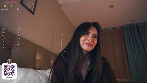 Media: Video of a smiling, fair-skinned woman with long black hair, wearing a black jacket, in a dimly lit bedroom with a bed, lamp, and curtains.