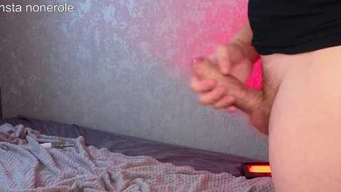 A video of a man's erect penis, partially obscured by his hand, standing on a bed with a grey blanket. The background features a textured, pale grey wall with pinkish-purple light.