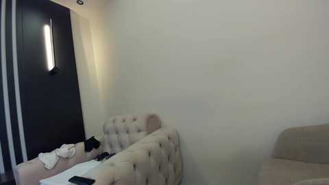 Media: Video of a minimalist bedroom corner with beige tufted headboard, white bedding, dark wall, and a vertical light fixture.