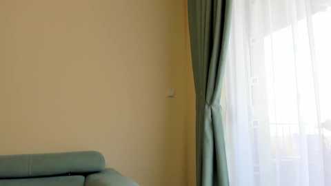 Media: Video of a minimalist room with a beige wall, a light blue sofa, and a window covered by sheer white curtains.