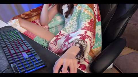 Media: Video of a woman with long brown hair wearing a colorful, patterned blanket and a black shirt, typing on a black mechanical keyboard. Background features a black office chair and a brown couch.