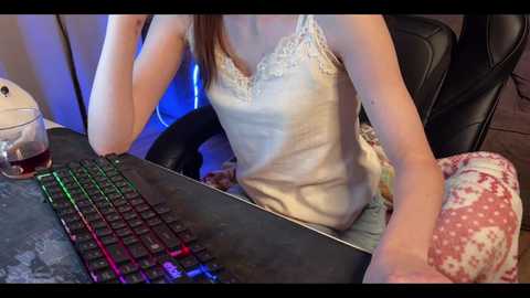 Media: A video of a young woman, wearing a white lace tank top and patterned pajama pants, typing on a black keyboard with colorful LED lights. She sits on a black leather chair in a dimly lit room with blue lighting.