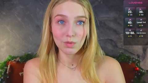 Media: Video of a young, fair-skinned woman with long blonde hair and blue eyes, wearing a delicate necklace. Background shows a green wreath and a dark stone wall.