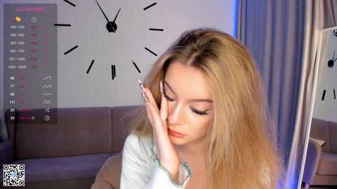 Media: Video of a young blonde woman with long hair, wearing makeup, sitting on a beige couch, holding her face in a distressed manner. Background features a white wall with a large clock and curtains.