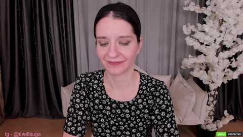 Media: Video of a smiling woman with light skin and dark hair, wearing a black floral dress, seated in a room with gray curtains and a white floral arrangement.