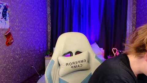 Media: Video of a person wearing a beige gaming chair with \"CAMEAR\" text, in a dimly lit room with purple lighting, Christmas stockings, and a heart-shaped neon sign.