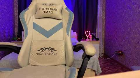 Media: Video of a gamer in a white and blue gaming chair with a tribal tattoo on the back, playing in a dimly lit room with purple walls and a heart-shaped neon light.