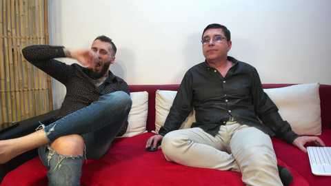 Media: Video of two middle-aged men with facial hair, one in a black shirt and jeans, the other in a black shirt and beige pants, sitting on a red couch in a modern living room.