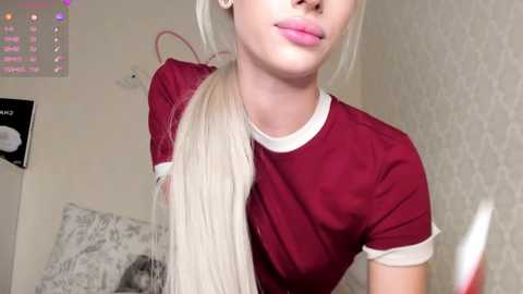 Media: Video of a fair-skinned woman with long blonde hair, wearing a maroon T-shirt, standing in a room with a white, patterned wall.