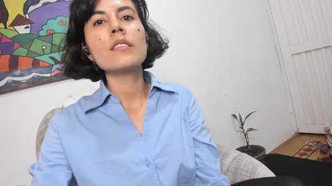 Media: Video of a young woman with short, black hair, wearing a light blue button-up shirt, sitting on a couch in a colorful, modern room with abstract art on the wall.