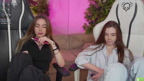 Media: Video of two young women, one with long brown hair in a black top, the other in a white hoodie, seated on gaming chairs. Background features pink lighting and green plants.