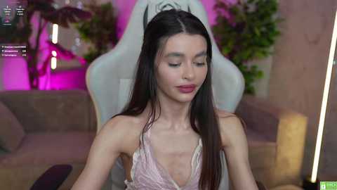 Media: A video of a young, slender woman with long, straight dark hair, wearing a low-cut, pink satin dress, sitting in a plush, white gaming chair. The background features a modern, dimly lit room with greenery and purple lighting.