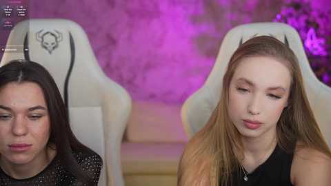 Media: Video of two young women seated in white gaming chairs with purple LED lights behind them. One woman, with long black hair, wears a black sequin top, and the other, with long blonde hair, wears a black top.