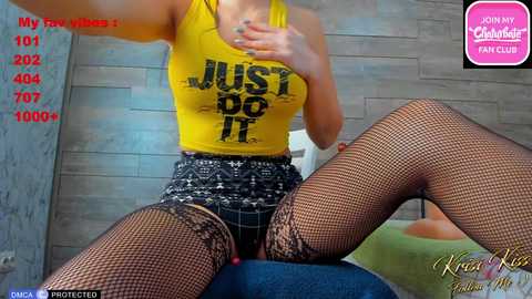 Media: Video of a curvaceous woman with light skin, wearing a yellow crop top, black lace panties, and fishnet stockings, seated on a blue cushion. Background features a wooden wall and a pink \"MyFreeCams\" watermark.