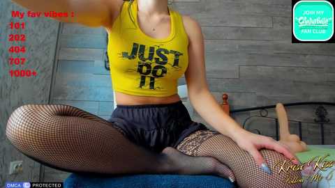 Media: Video of a fair-skinned woman with medium breasts, wearing a yellow crop top, fishnet stockings, and black shorts, sitting on a bed. Background shows a wooden wall, a phone screen with text, and a sex toy.