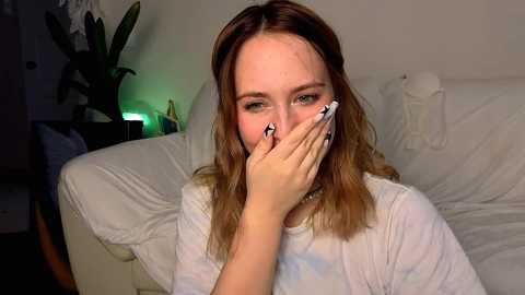 Media: Video of a young woman with shoulder-length brown hair, wearing a white T-shirt, covering her mouth with her hand, sitting on a white couch in a dimly lit room with a green lamp and a potted plant in the background.