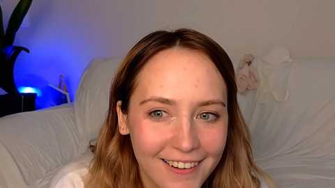 Media: Video of a smiling, light-skinned woman with shoulder-length, wavy, light brown hair, wearing a white top. She sits on a white bed with a plush toy, against a dimly lit, white wall.