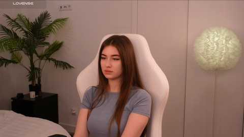 Media: Video of a young woman with long brown hair, wearing a light blue t-shirt, sitting in a white gaming chair in a minimalistic bedroom with a potted plant and a textured lamp.