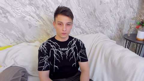 Media: A young man with short brown hair and fair skin sits on a bed in a dimly lit room. He wears a black T-shirt with white lightning bolts and grey pants. The background features textured white walls and a side table with a potted plant.