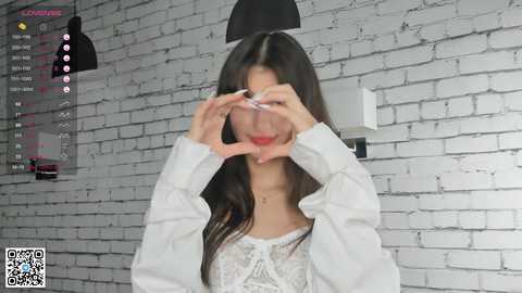 Media: Video of a young Asian woman with long dark hair, wearing a white lace top, forming a heart shape with her hands over her face. Background features a white brick wall, a black lamp, and a QR code.