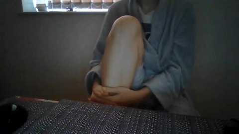 Media: Video of a person with light skin and short, light brown hair, wearing a gray sweatshirt, sitting on a patterned black and white blanket, holding a prosthetic leg. Background shows a window with closed blinds.