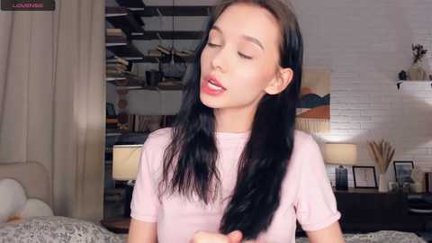 Media: A video of a young Asian woman with long black hair, wearing a pink T-shirt, leaning against a white brick wall. The room features a bed, a lamp, and a shelf with books and decorative items.