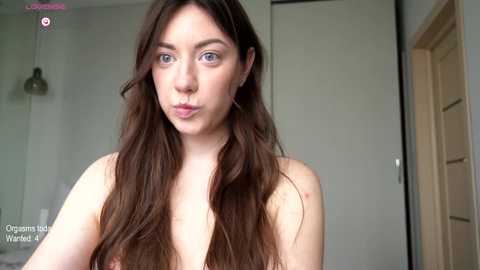 Media: Video of a young, fair-skinned woman with long brown hair, blue eyes, and no top, puckering her lips in a playful manner. Background shows a beige wardrobe and a minimalist room with soft lighting.