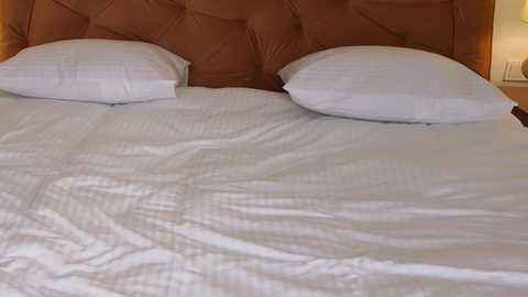 Media: Video of a neatly made bed with white sheets, two white pillows, and a tufted brown headboard in a modern, minimalist bedroom.