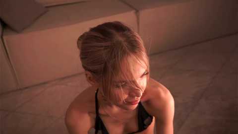 Media: A video of a young woman with fair skin, blonde hair, and a black top, sitting on a tiled floor. The setting is a modern, minimalistic room with beige walls and a cream-colored couch in the background.