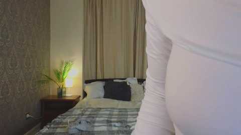 Media: Video of a cozy bedroom with beige curtains, a dark wooden headboard, white bedspread, and a potted plant beside a lamp on a nightstand.