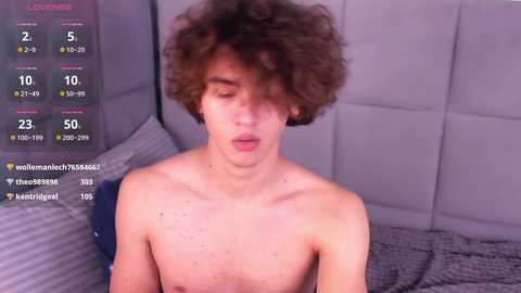 Media: A video of a young, fair-skinned, shirtless man with curly brown hair, lying on a bed with gray bedding, looking expressionless.