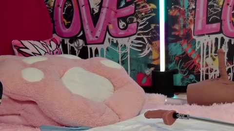 Media: Video of a plush, pink, polka-dotted dog on a soft, pink rug in a room with graffiti-covered walls, featuring the word \"LOVE.\