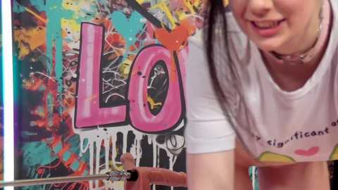 Media: Video of a smiling young woman with dark hair wearing a white t-shirt, leaning against a vibrant, colorful graffiti wall with the word \"LOVE\" in large, pink letters.