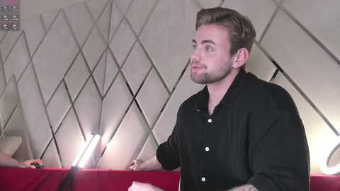 Media: Video of a young man with light brown hair and a beard, wearing a black shirt, sitting on a red sofa. The background features geometric diamond-patterned wall tiles and bright lighting.