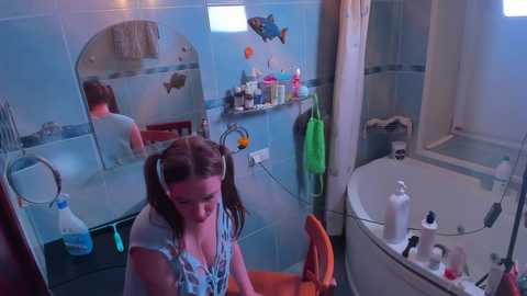 Media: Video of a young girl with pigtails in a bathroom with blue tiles, a mirror, a white bathtub, and toiletries.