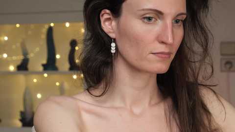 Media: Video of a woman with long, dark hair, light skin, and green eyes, wearing elegant dangling earrings, standing indoors with a blurred background featuring a warm, yellow-lit shelf.