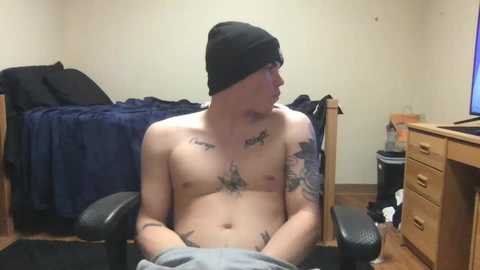 Media: Video of a shirtless, muscular, light-skinned man with tattoos on his chest and arms, wearing a black beanie, sitting in a dark chair, in a messy bedroom with a wooden dresser and a bed with blue sheets.