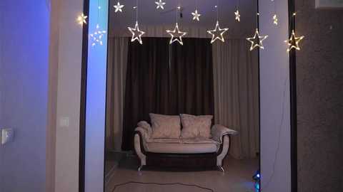 Media: A video of a cozy, modern living room with a beige sofa, star-shaped string lights hanging above, and dark curtains in the background.