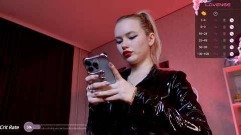 Media: Video of a fair-skinned woman with a ponytail, wearing black latex, taking a selfie. Background shows dimly lit room with red lighting.