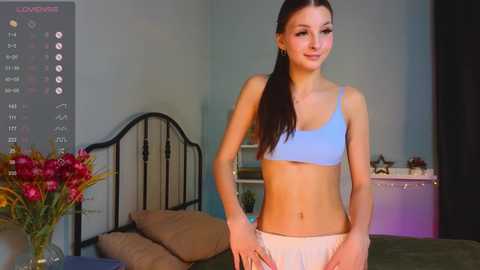 Media: Video of a slender, young woman with fair skin, long brown hair, wearing a light blue sports bra and white shorts, standing in a pastel-colored bedroom with a black metal bed frame, floral arrangement, and soft lighting.