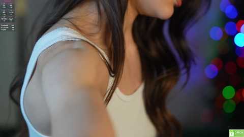 Media: A close-up video of a woman with long, wavy brown hair, wearing a white tank top, her side profile in focus against a colorful, blurred bokeh background.