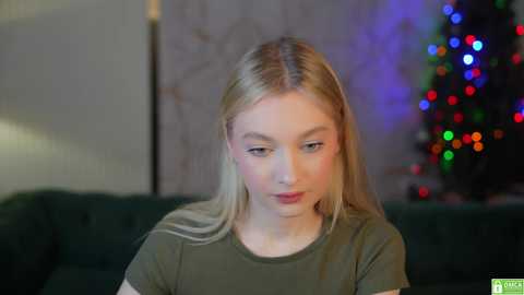 Media: Video of a young, fair-skinned, blonde woman with straight hair, wearing a green t-shirt, seated indoors. Background features a blurred Christmas tree with multicolored lights.