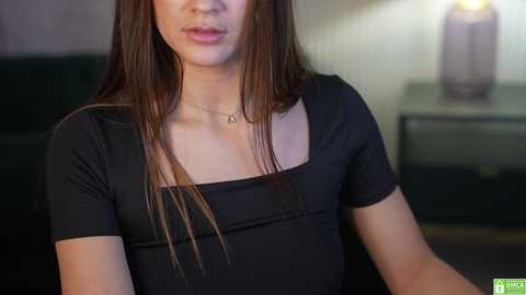 Media: Video of an Asian woman with long, straight brown hair, wearing a black, short-sleeved top with a square neckline, sitting indoors. Background features a blurred dark couch and a lamp on a side table.