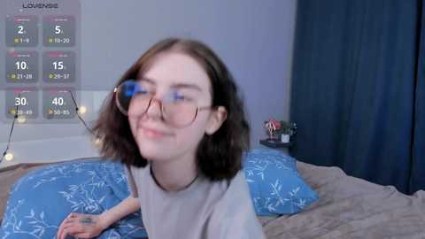 Media: Video of a young woman with shoulder-length brown hair, wearing glasses, smiling, in a cozy bedroom with a blue-patterned duvet, a calendar, and a dark curtain.